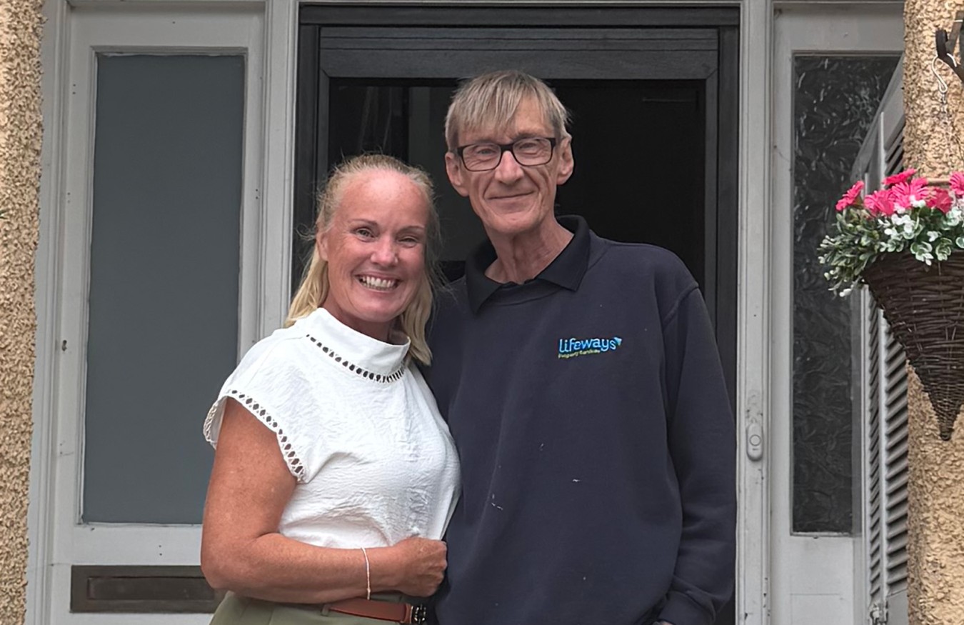 Smiling couple standing close together in doorway, woman in white top, man in dark polo with Lifeways logo.