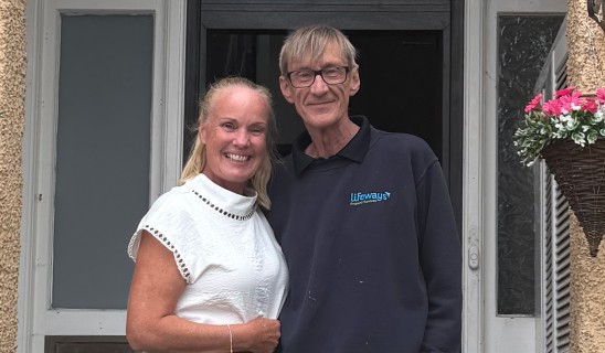 Smiling couple standing close together in doorway, woman in white top, man in dark polo with Lifeways logo.