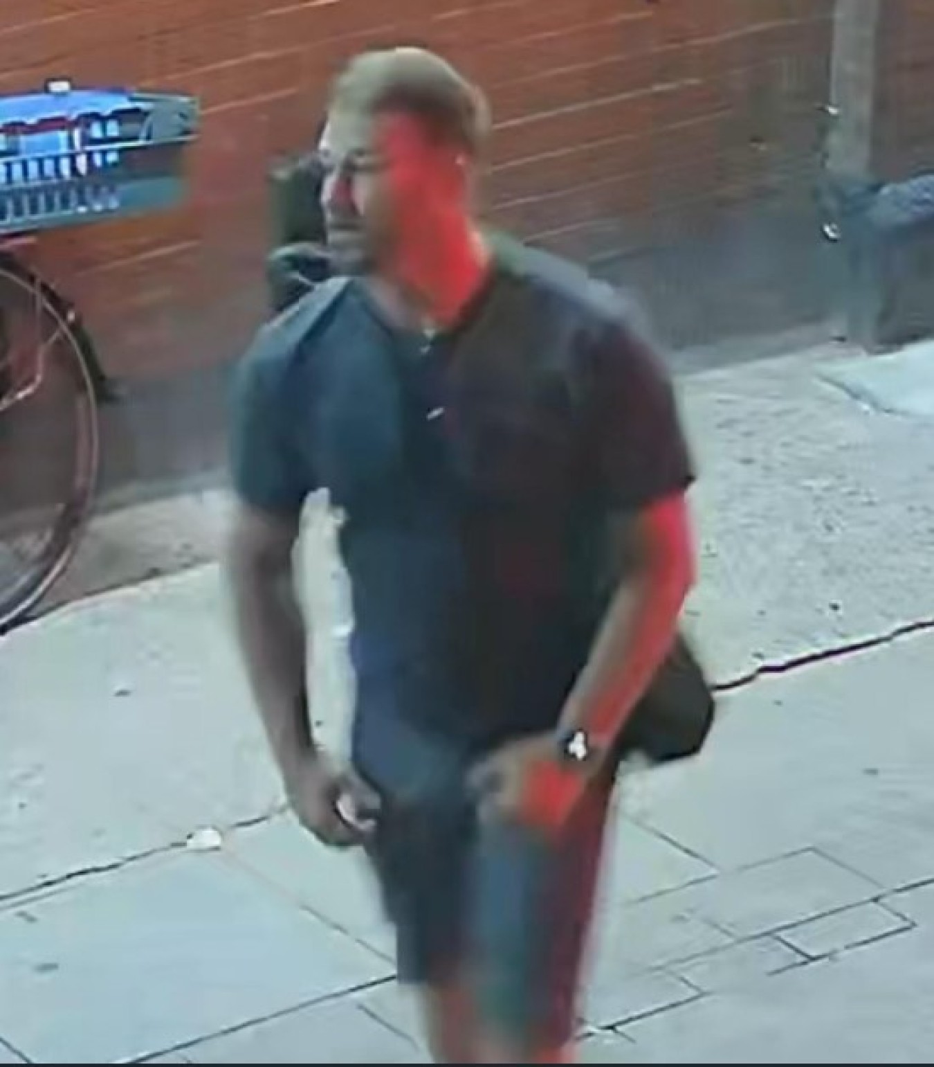 Man in dark t-shirt and shorts walking along a sidewalk carrying a bag.