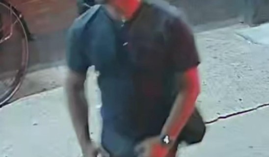 Man in dark t-shirt and shorts walking along a sidewalk carrying a bag.