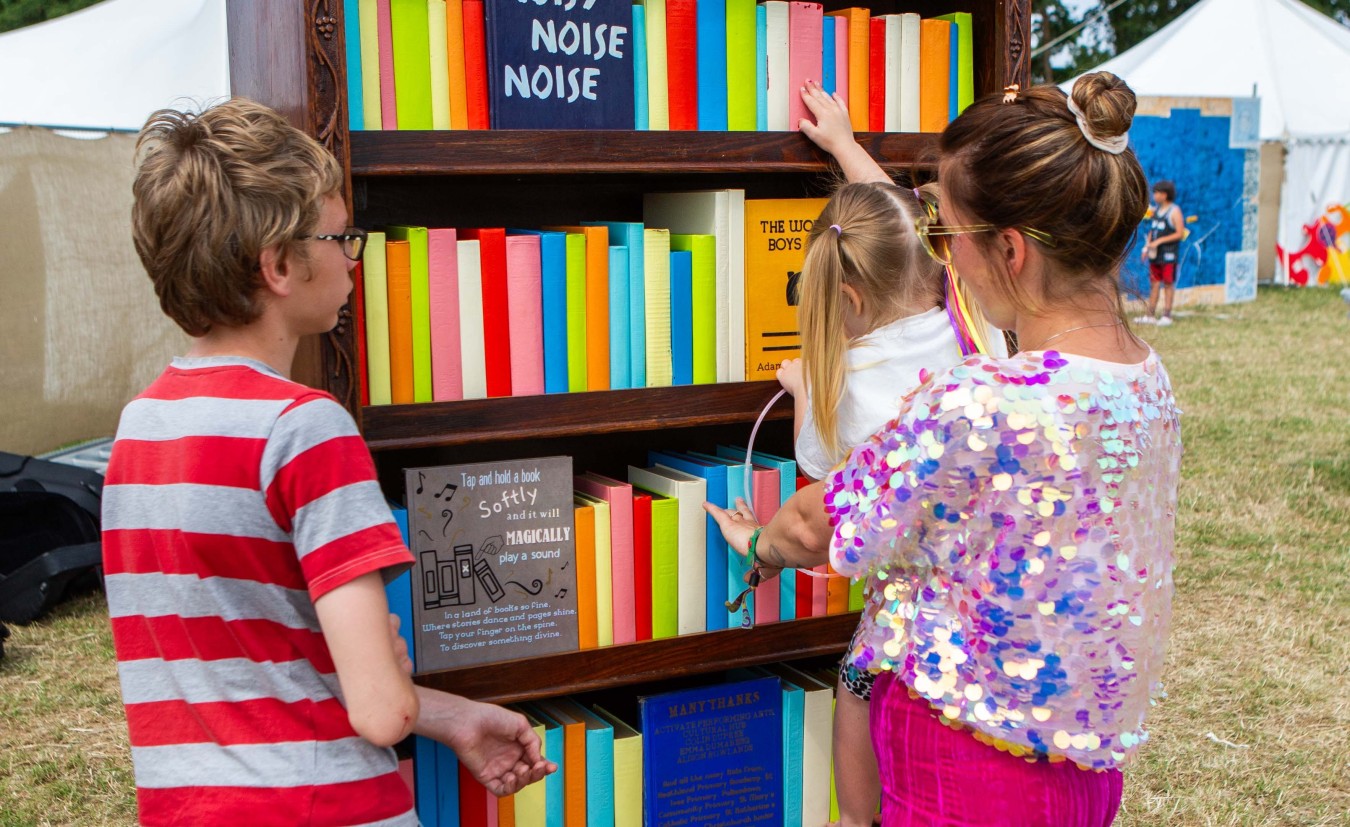 Children browse a bright, rainbow-colored bookshelf outdoors at an event.