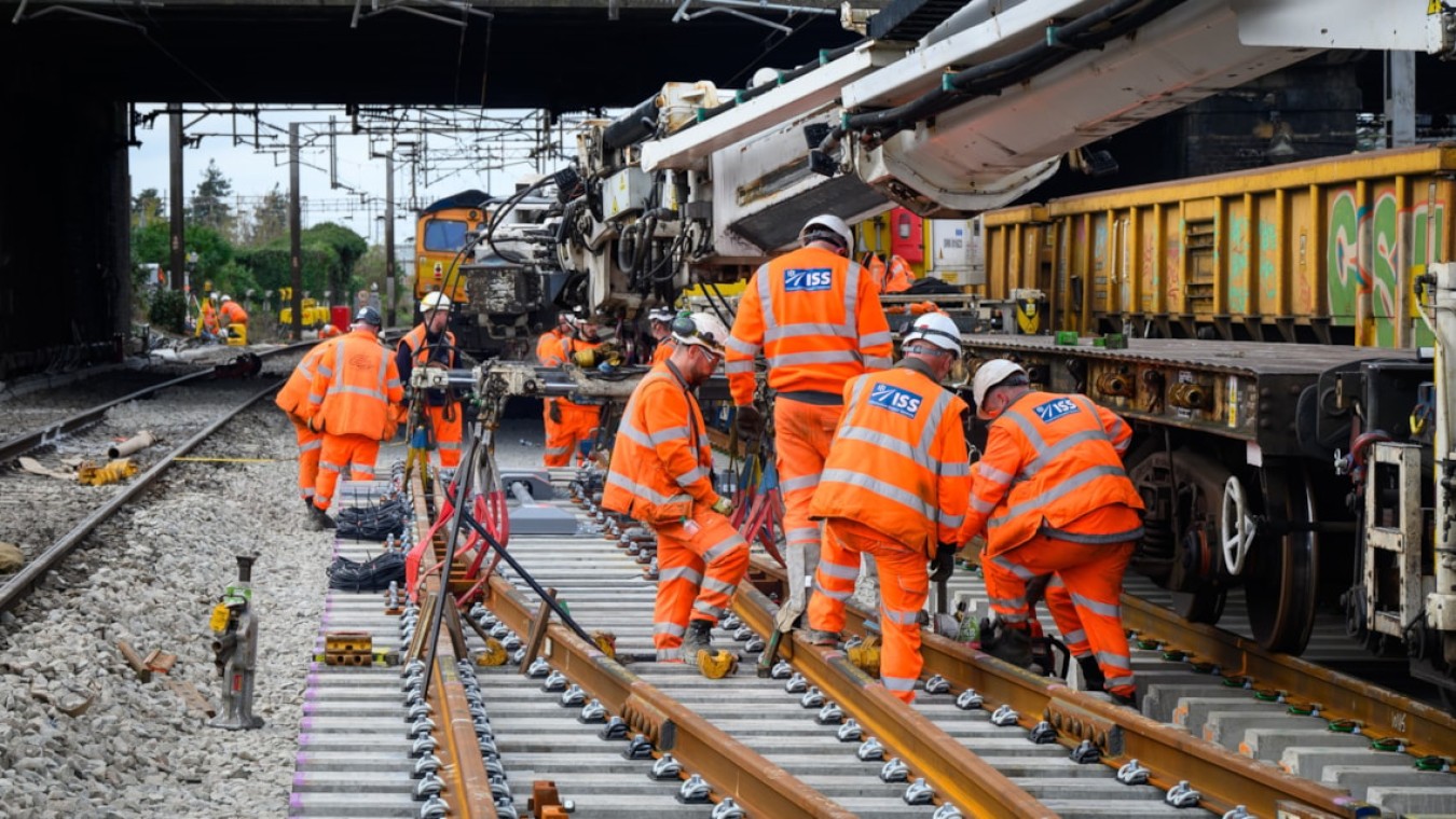 Railway workers in bright orange overalls assembling track under a bridge, large machinery and a freight train nearby.