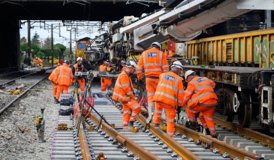 Railway workers in bright orange overalls assembling track under a bridge, large machinery and a freight train nearby.
