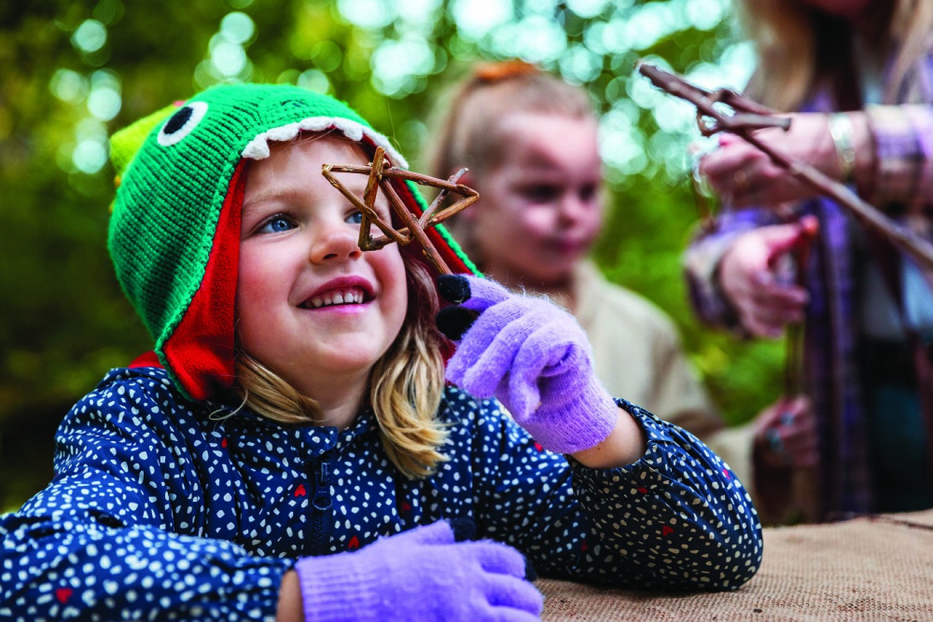 Child wearing a green knitted hat with eye design, smiling while holding a twig star.
