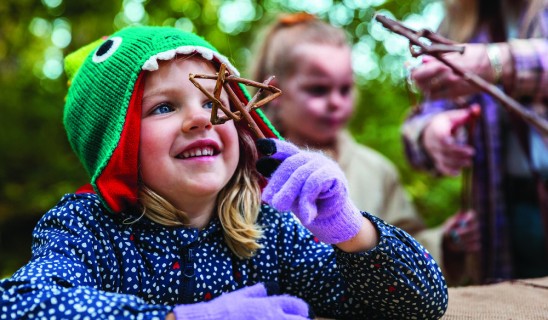 Child wearing a green knitted hat with eye design, smiling while holding a twig star.