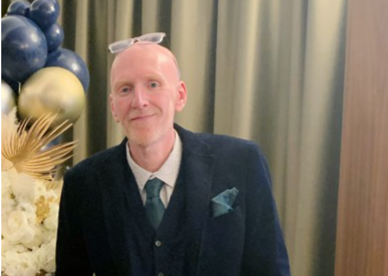 Man in dark suit with teal tie and pocket square at formal event, background balloons and curtains