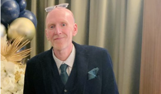 Man in dark suit with teal tie and pocket square at formal event, background balloons and curtains