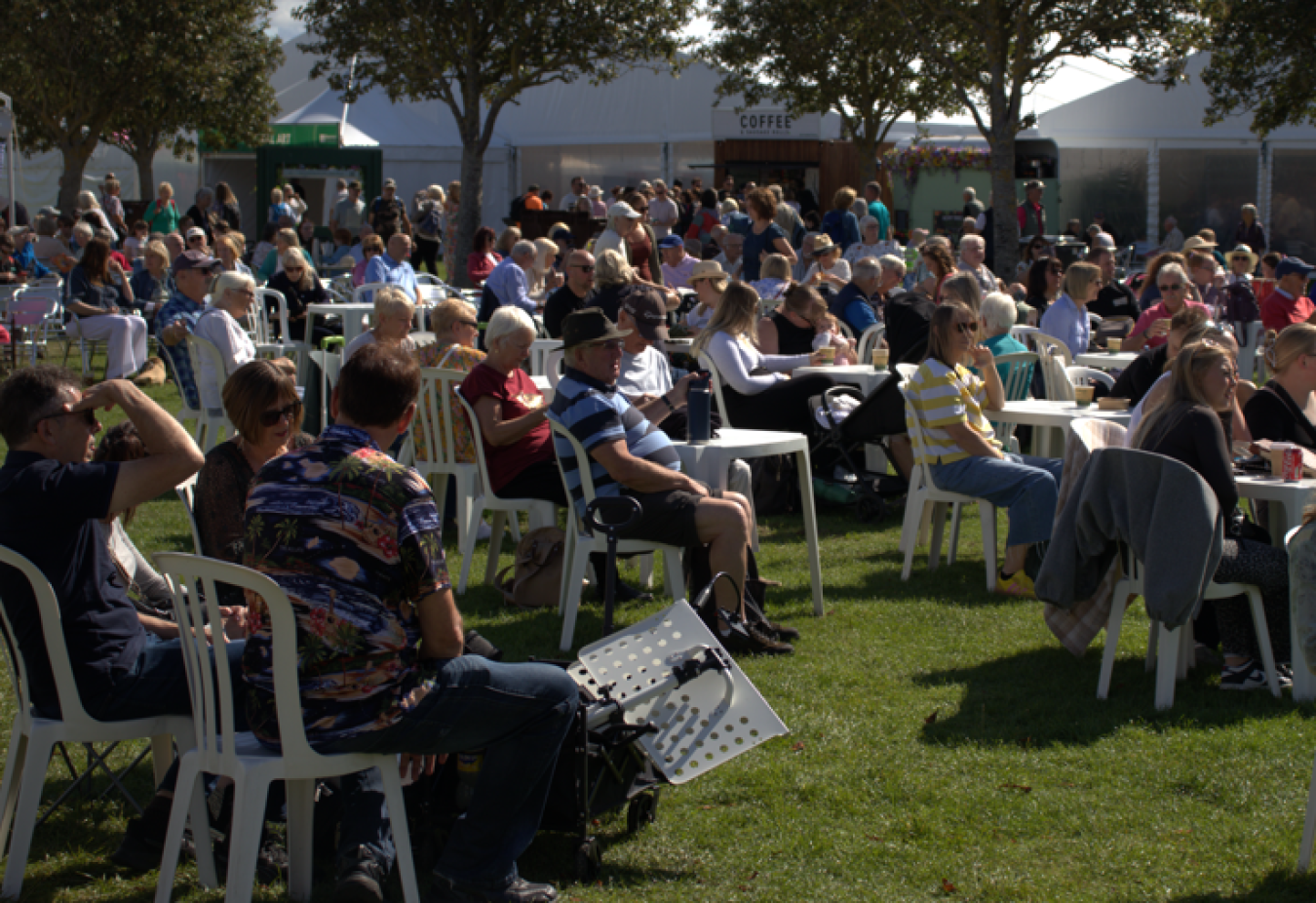 A crowd seated on white plastic chairs at an outdoor event under trees with a large white tent in the background.