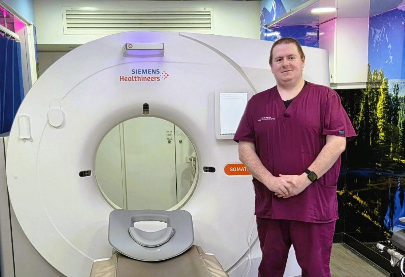 Healthcare worker in maroon scrubs standing beside a Siemens Somatom CT scanner in a medical facility.