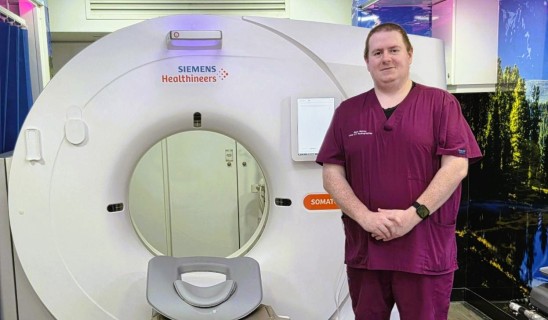 Healthcare worker in maroon scrubs standing beside a Siemens Somatom CT scanner in a medical facility.