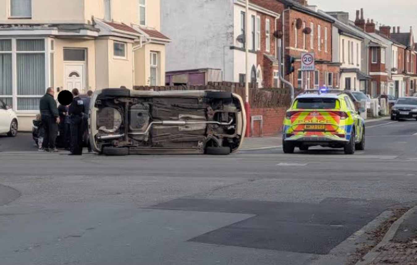 Car overturned on its side blocking residential street; police car and officers at scene with bystanders nearby.