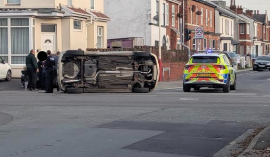 Car overturned on its side blocking residential street; police car and officers at scene with bystanders nearby.