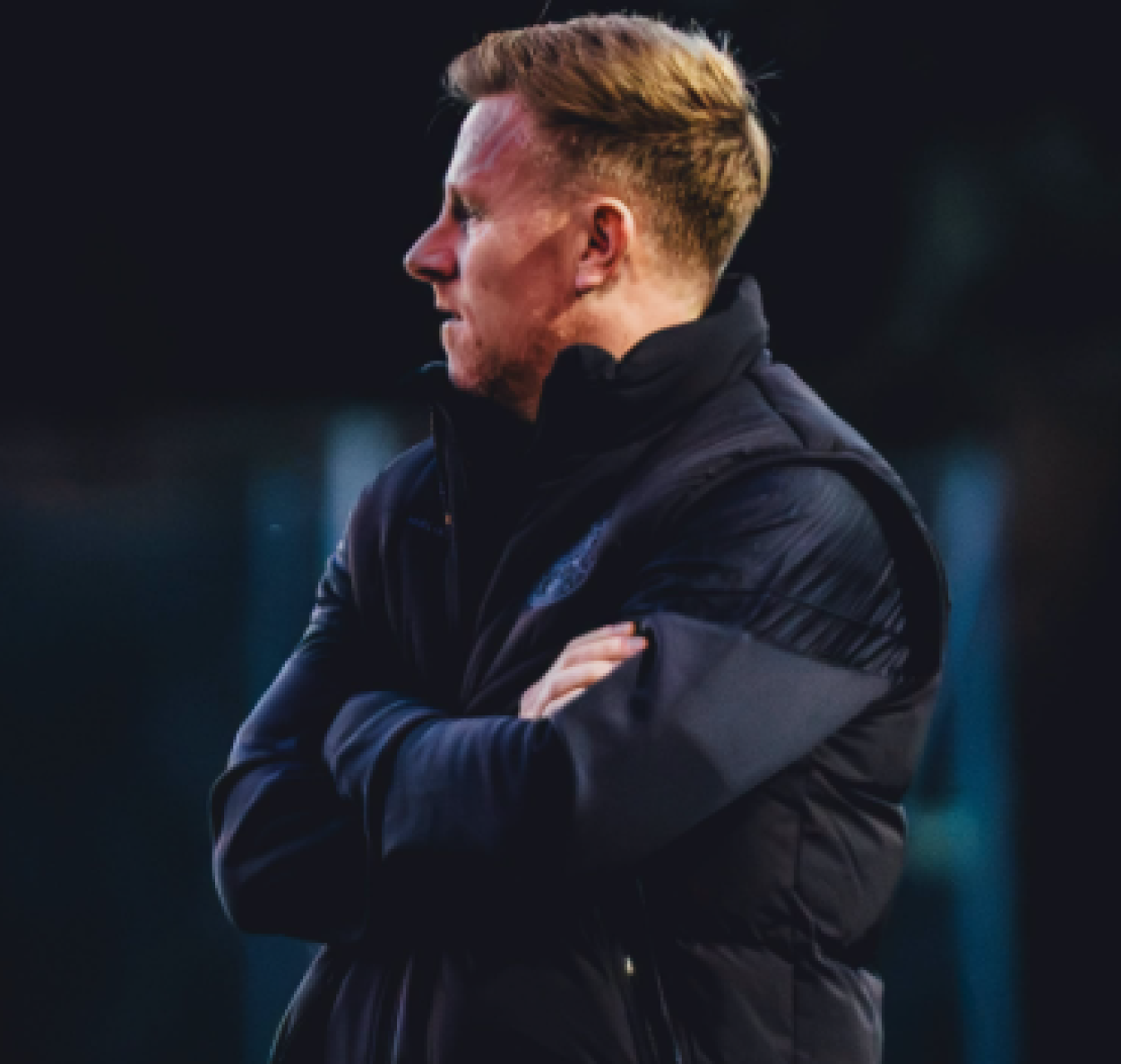 Coach standing in dark jacket with arms crossed, looking to the left