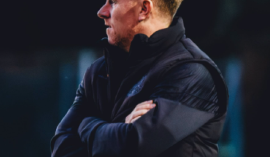 Coach standing in dark jacket with arms crossed, looking to the left