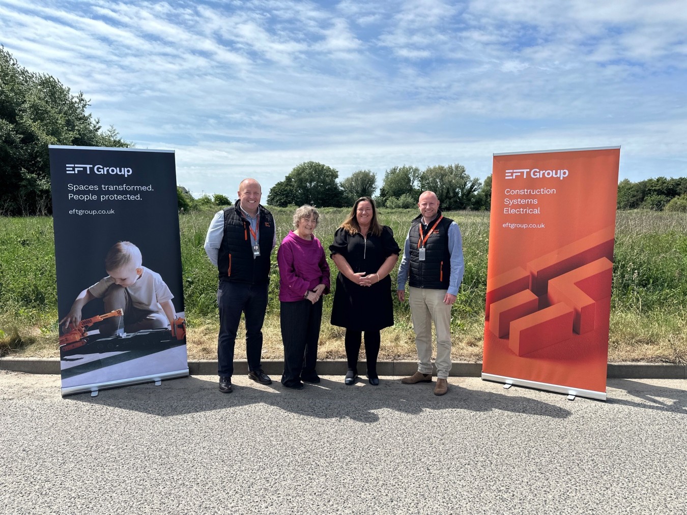 Four people standing outdoors between two banners for EFT Group, blue sky and trees behind.
