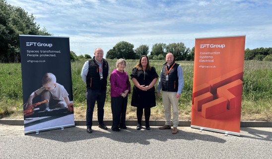 Four people standing outdoors between two banners for EFT Group, blue sky and trees behind.
