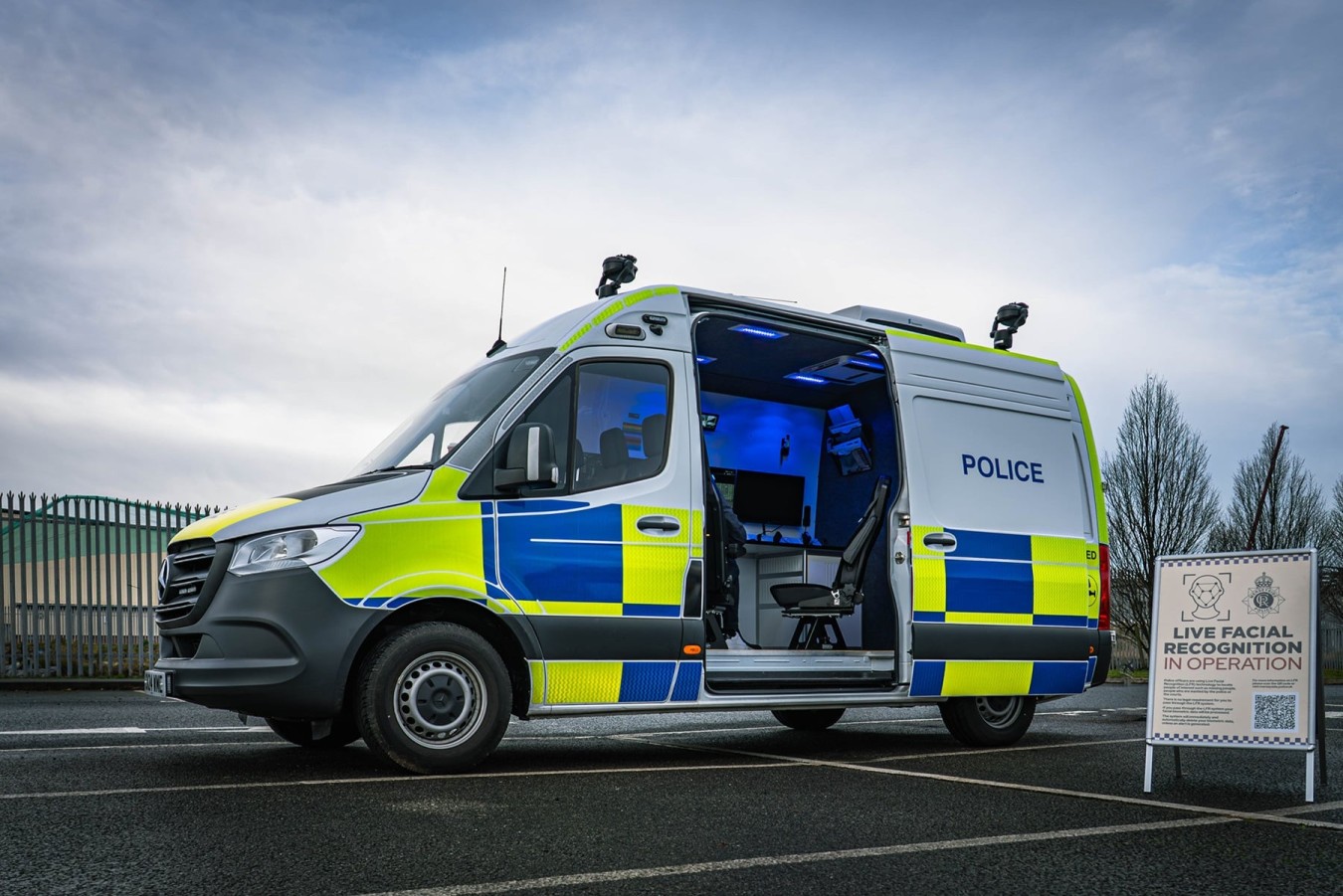 police van with open side door in a parking lot, blue lights inside, signboard about facial recognition nearby
