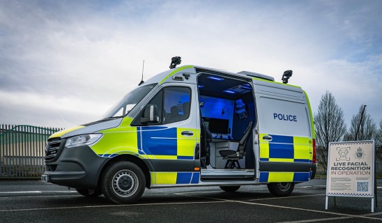 police van with open side door in a parking lot, blue lights inside, signboard about facial recognition nearby