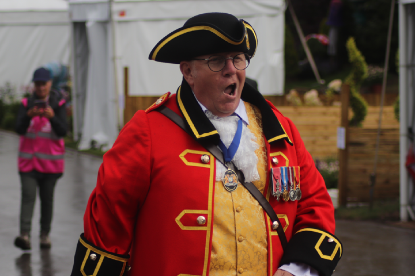 Man in red ceremonial coat and hat shouting, medals on chest, outdoors at event.