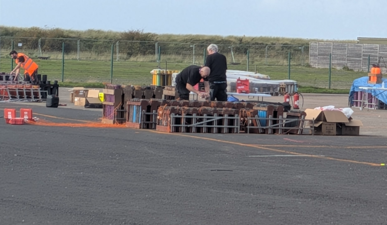 Two workers near pallets of bricks and crates at an open tarmac area, with fenced boundary and blue tarps in the background.