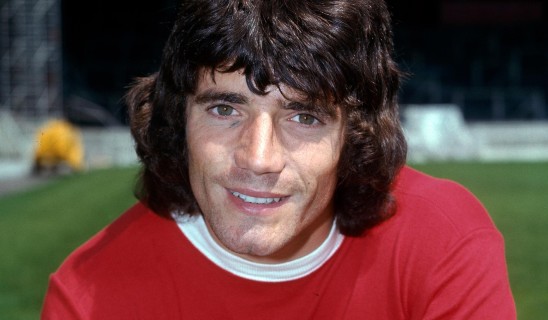 Man with dark hair smiling, wearing red shirt, outdoors at night.