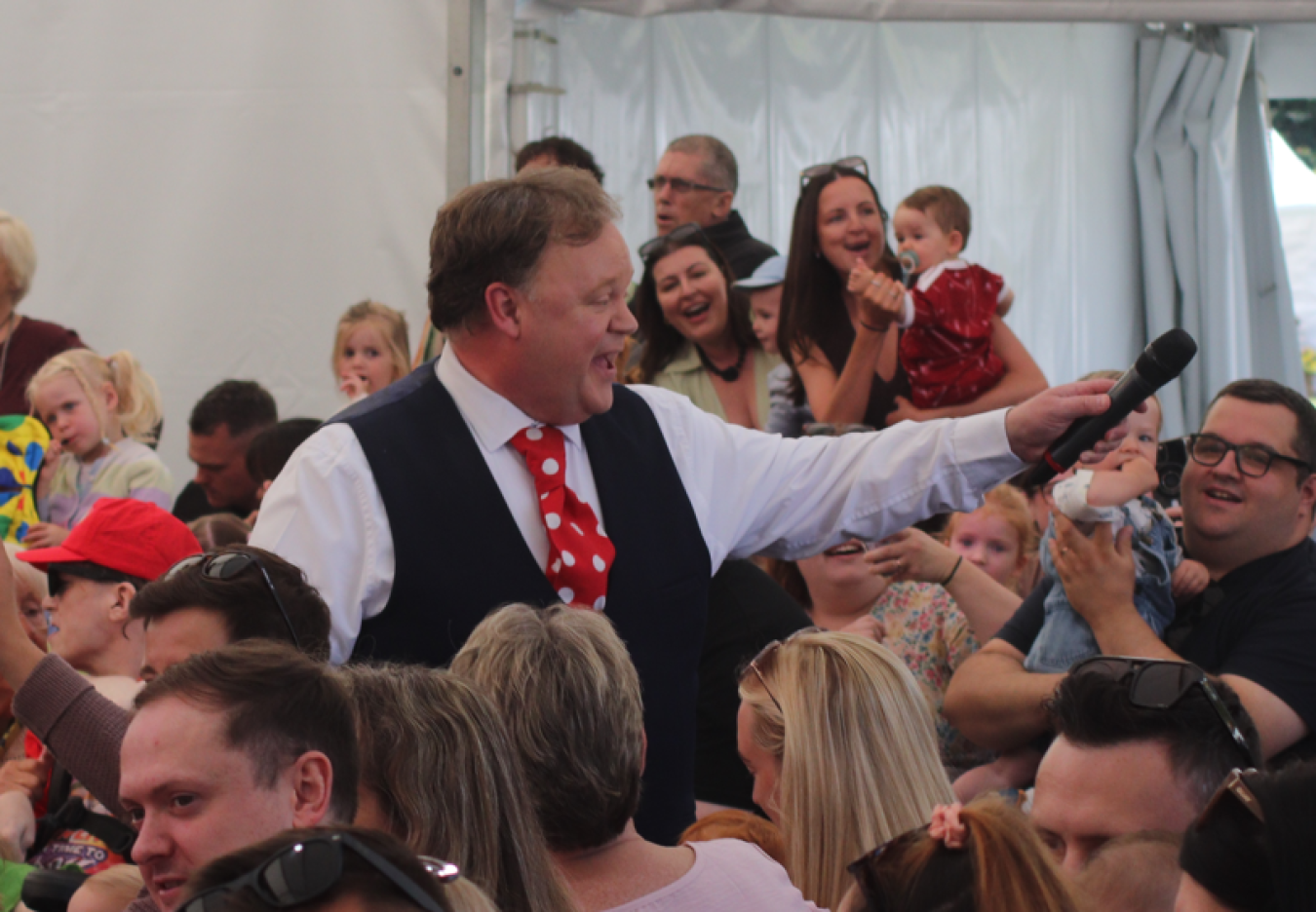 Entertainer with microphone addressing a crowded indoor event, many spectators smiling and reaching out.