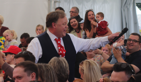 Entertainer with microphone addressing a crowded indoor event, many spectators smiling and reaching out.