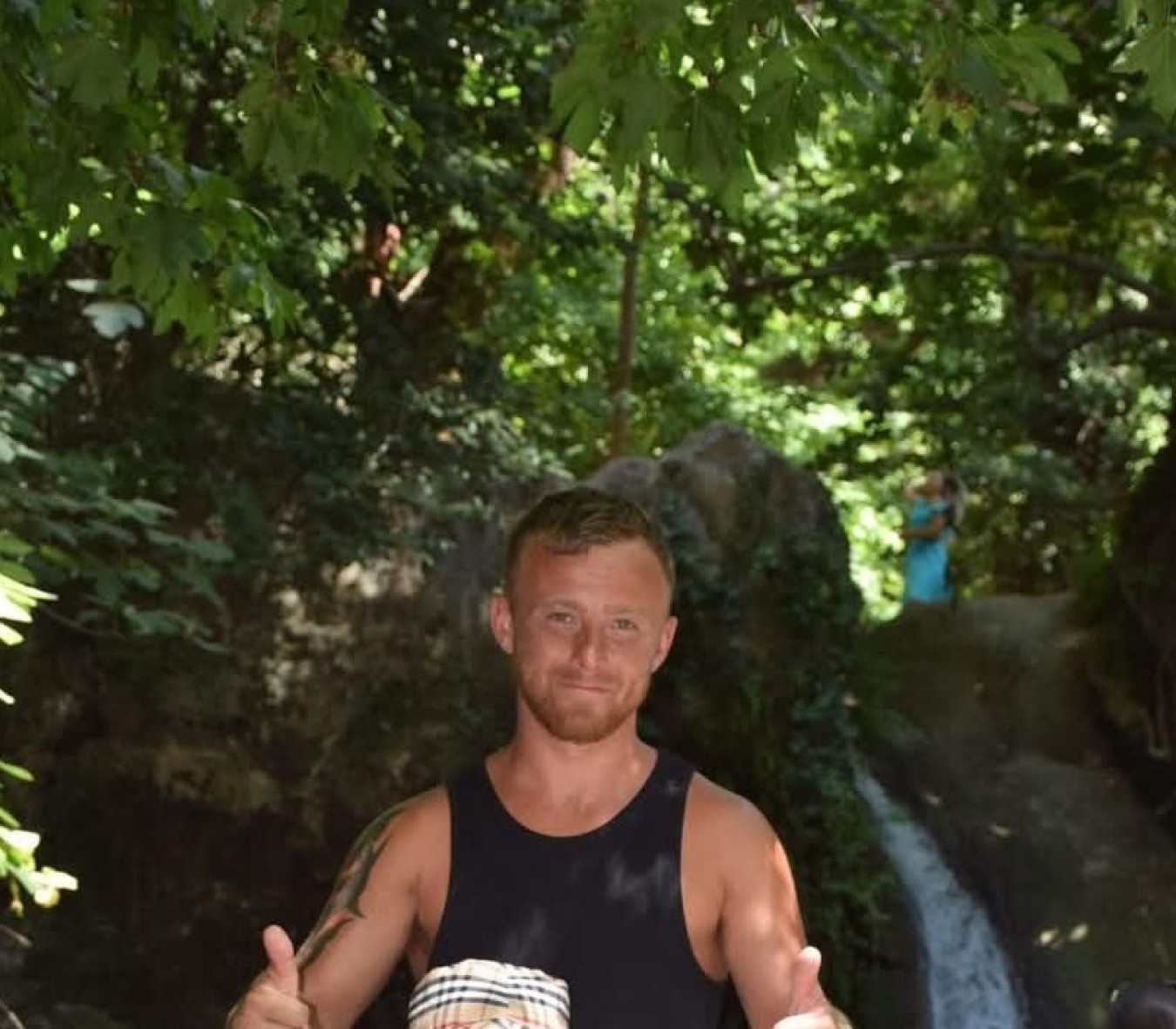 Man in sleeveless black shirt giving thumbs up near a waterfall in a green woodland setting.
