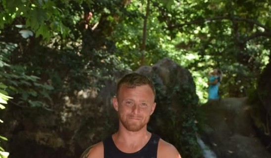 Man in sleeveless black shirt giving thumbs up near a waterfall in a green woodland setting.
