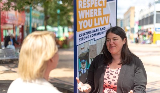 Two women talking on a sunny street, activist banner saying 'Respect Where You Lviv' in background.