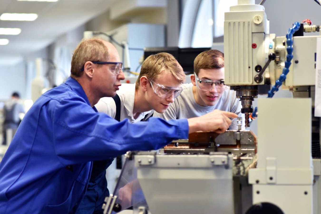 Three workers wearing safety goggles operate a large industrial machine in a bright workshop.