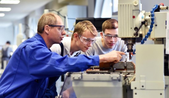 Three workers wearing safety goggles operate a large industrial machine in a bright workshop.