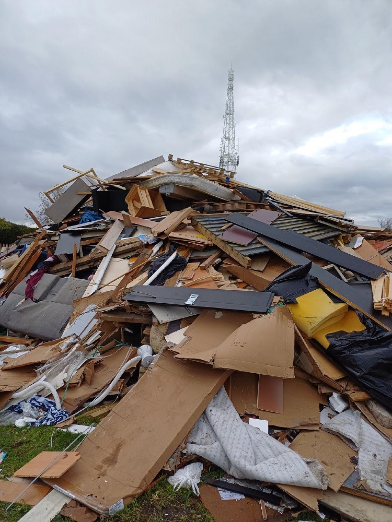 Piles of demolished building debris and rubble strewn across a field with a tall antenna tower in the background under a cloudy sky.