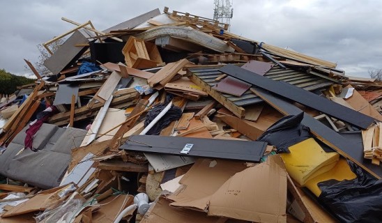 Piles of demolished building debris and rubble strewn across a field with a tall antenna tower in the background under a cloudy sky.