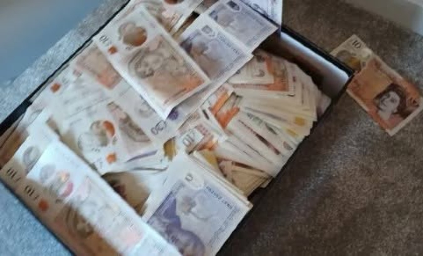 Stacks of banknotes scattered in a wooden drawer or bag on a carpeted floor.
