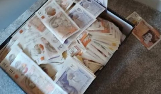 Stacks of banknotes scattered in a wooden drawer or bag on a carpeted floor.