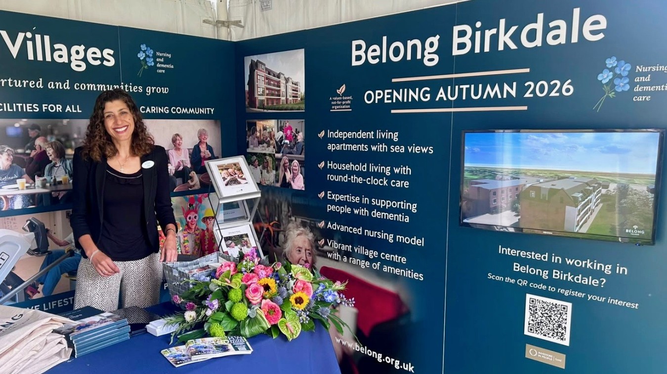 Smiling woman standing at a blue promotional display for Belong Birkdale autumn 2026 with a bouquet of flowers on the table.
