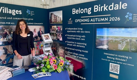 Smiling woman standing at a blue promotional display for Belong Birkdale autumn 2026 with a bouquet of flowers on the table.