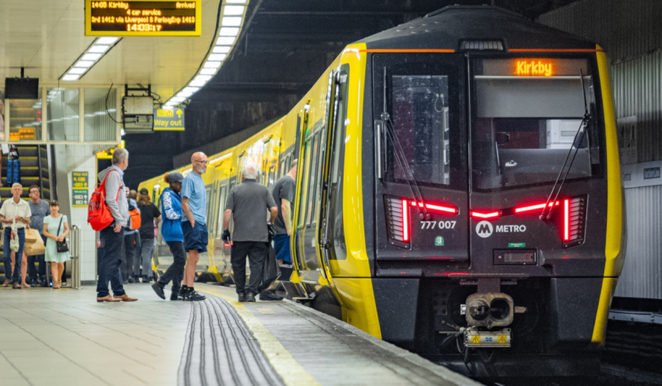 Commuters boarding a yellow and black London Metro train at a platform, with a digital display showing destination Kirkby.