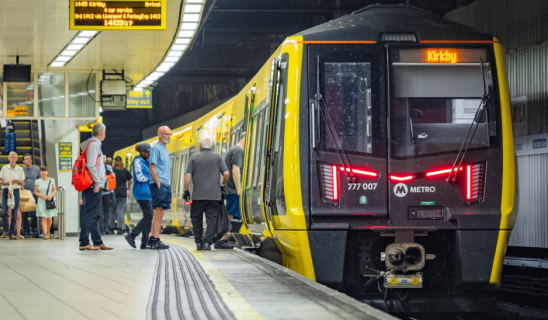Commuters boarding a yellow and black London Metro train at a platform, with a digital display showing destination Kirkby.